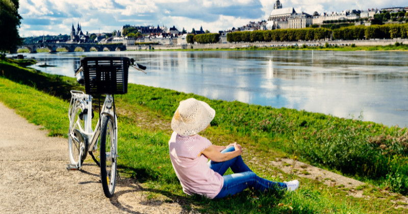 Woman sits peacefully by the river in France, taking a break next to her bicycle